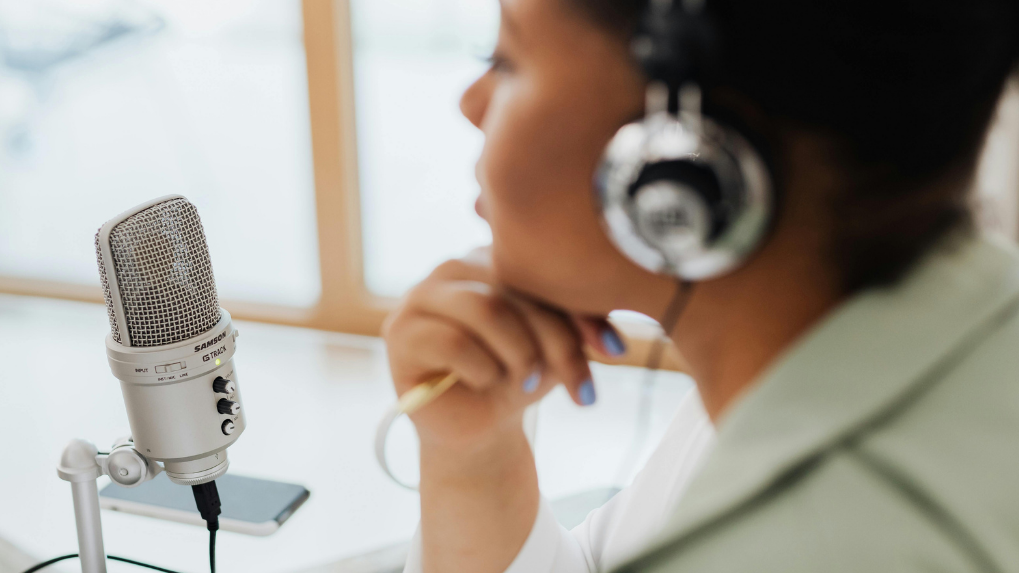 A woman wearing over-ear headphones sits at a desk, positioned beside a microphone on a stand. They are recording a podcast. 