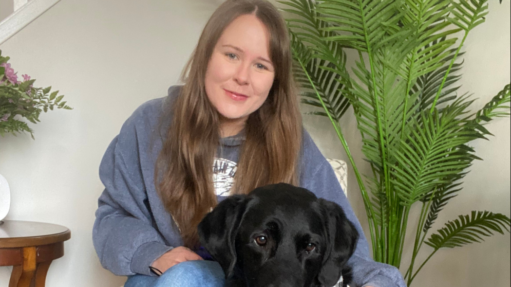 Taylor, a young woman with long brown hair, sits in a chair with her arm around her guide dog Wallace, a black lab wearing a CNIB harness, who sits beside her.