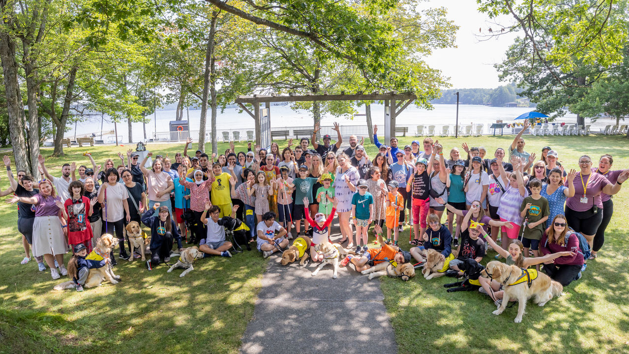 A large group of people of all ages, including many who are accompanied by buddy dogs or holding white canes, raise their hands in the air joyfully at Lake Joe - with the lake visible behind them.