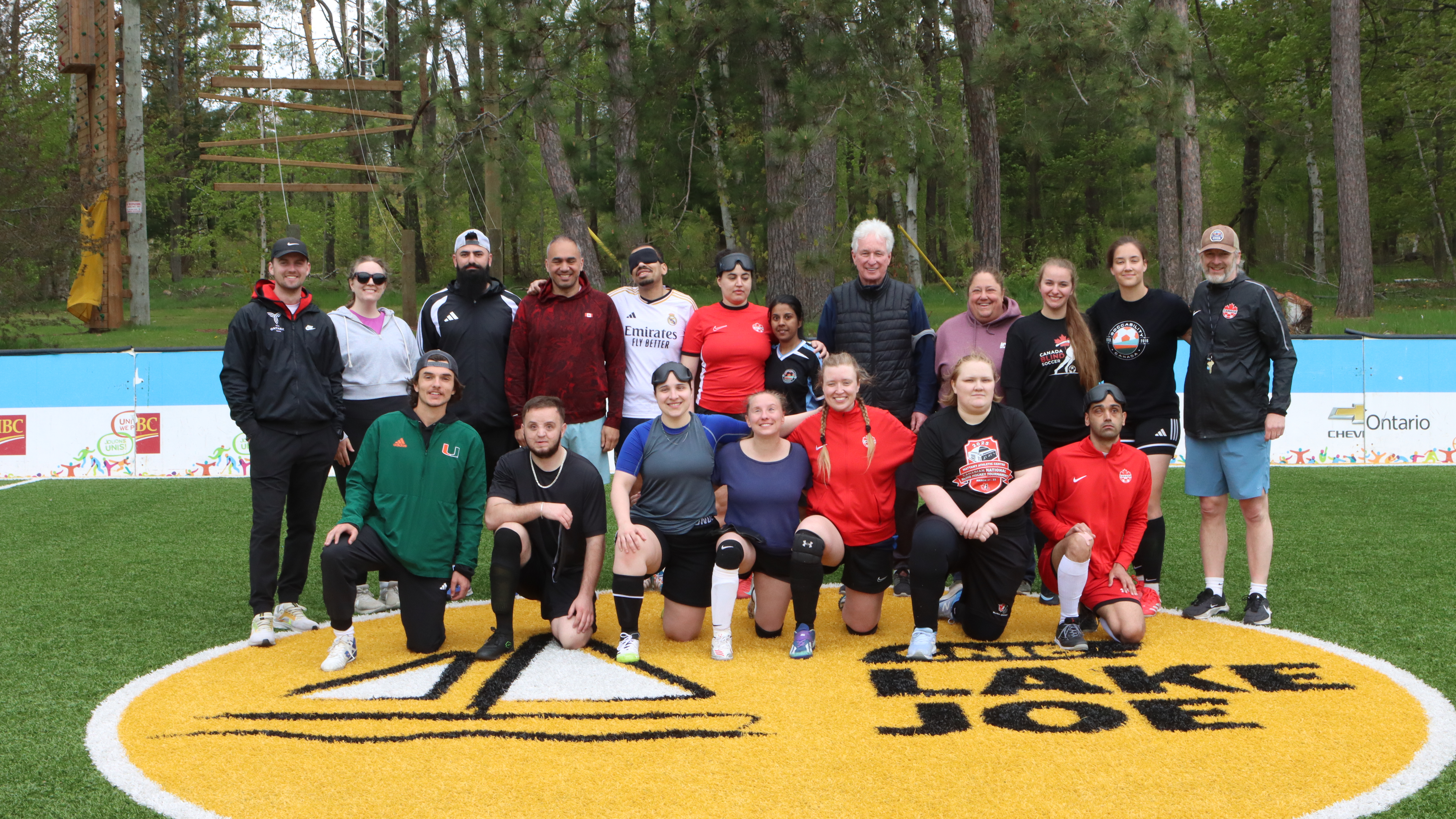 A group of blind soccer players of varying heights, ages, and skin tones stand in athletic gear on the accessible soccer pitch at CNIB Lake Joe, with a blind soccer ball in front of them.