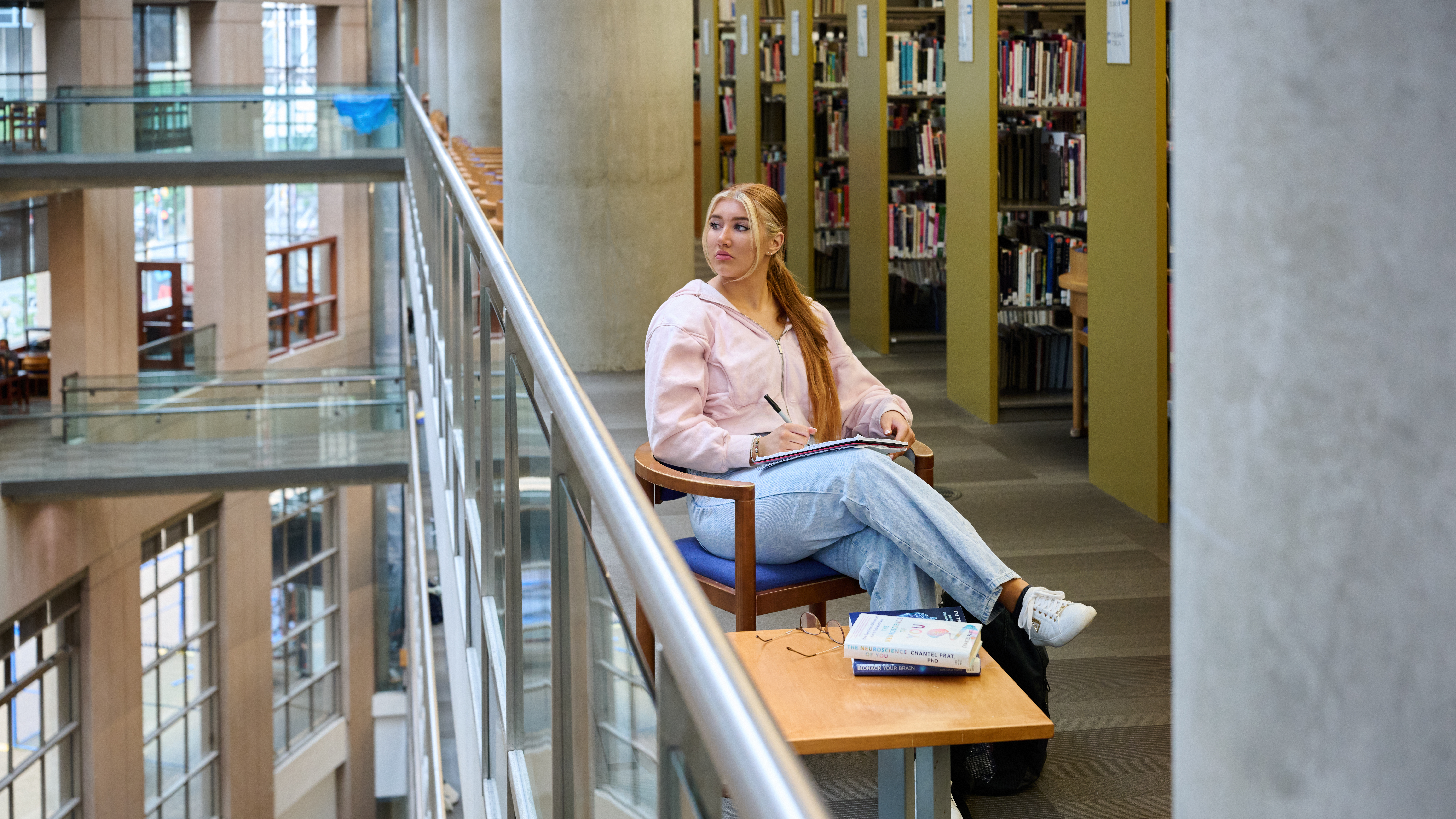 Ashlyn, wearing a pink sweater and jeans, sits in a wooden chair beside the glass railing of a multi floored library. She has a notebook and pen on her lap and is looking out over the railing's edge.