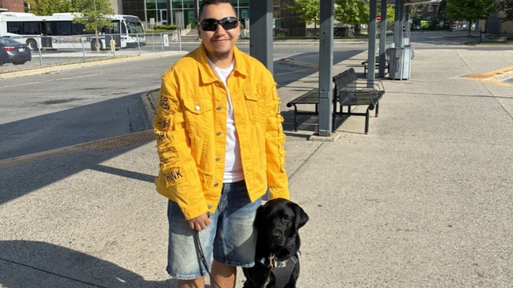 Tye stands beside his black Labrador guide dog, Zed, at an outdoor transit station. Tye hold’s Zed’s harness and leash. 