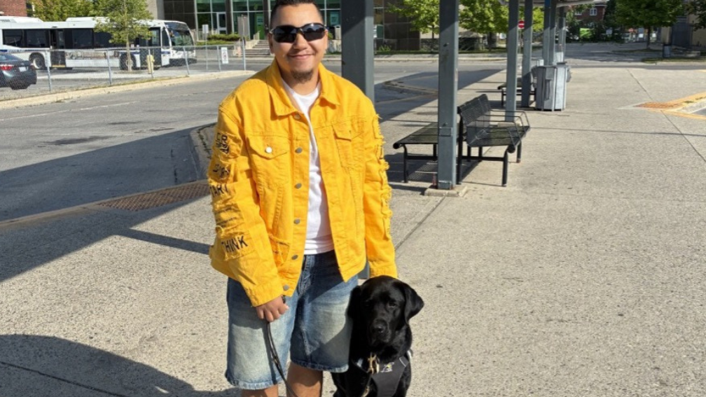 Tye stands beside his black Labrador guide dog, Zed, at an outdoor transit station. Tye hold’s Zed’s harness and leash. 