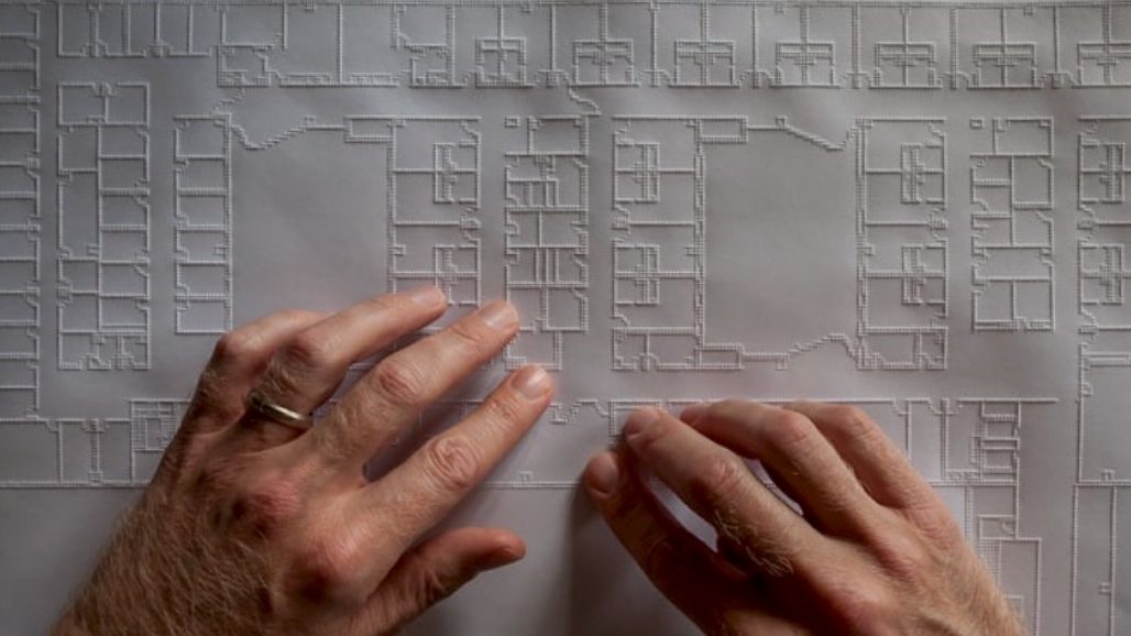Two hands move across a large tactile architectural floor plan. Raised lines outline rooms, corridors, walls, and other features of the building layout.