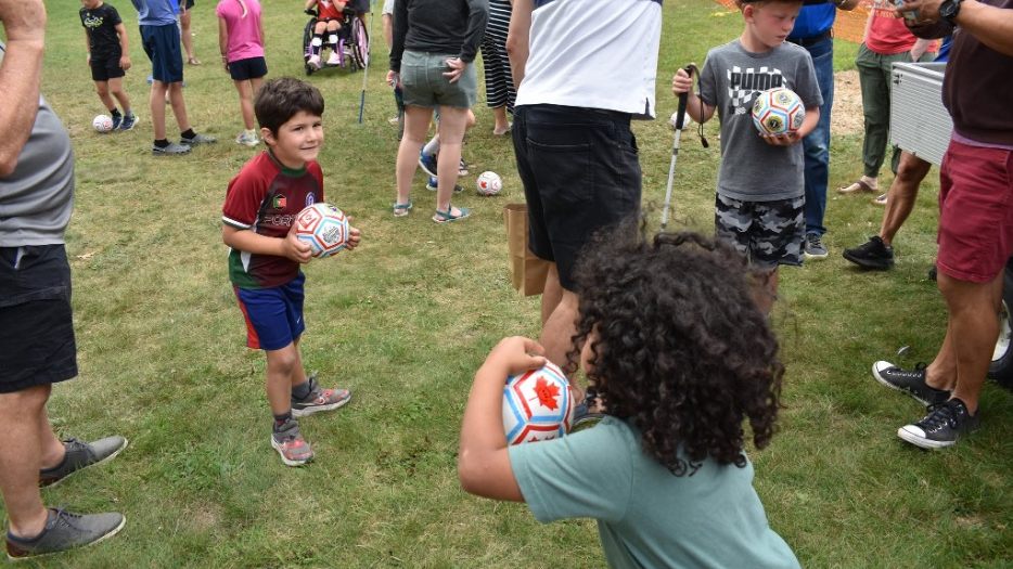 A crowd of children and adults gather on a grassy lawn for a soccer activity. Many of them are holding small white soccer balls in their hands or kicking them around their feet.