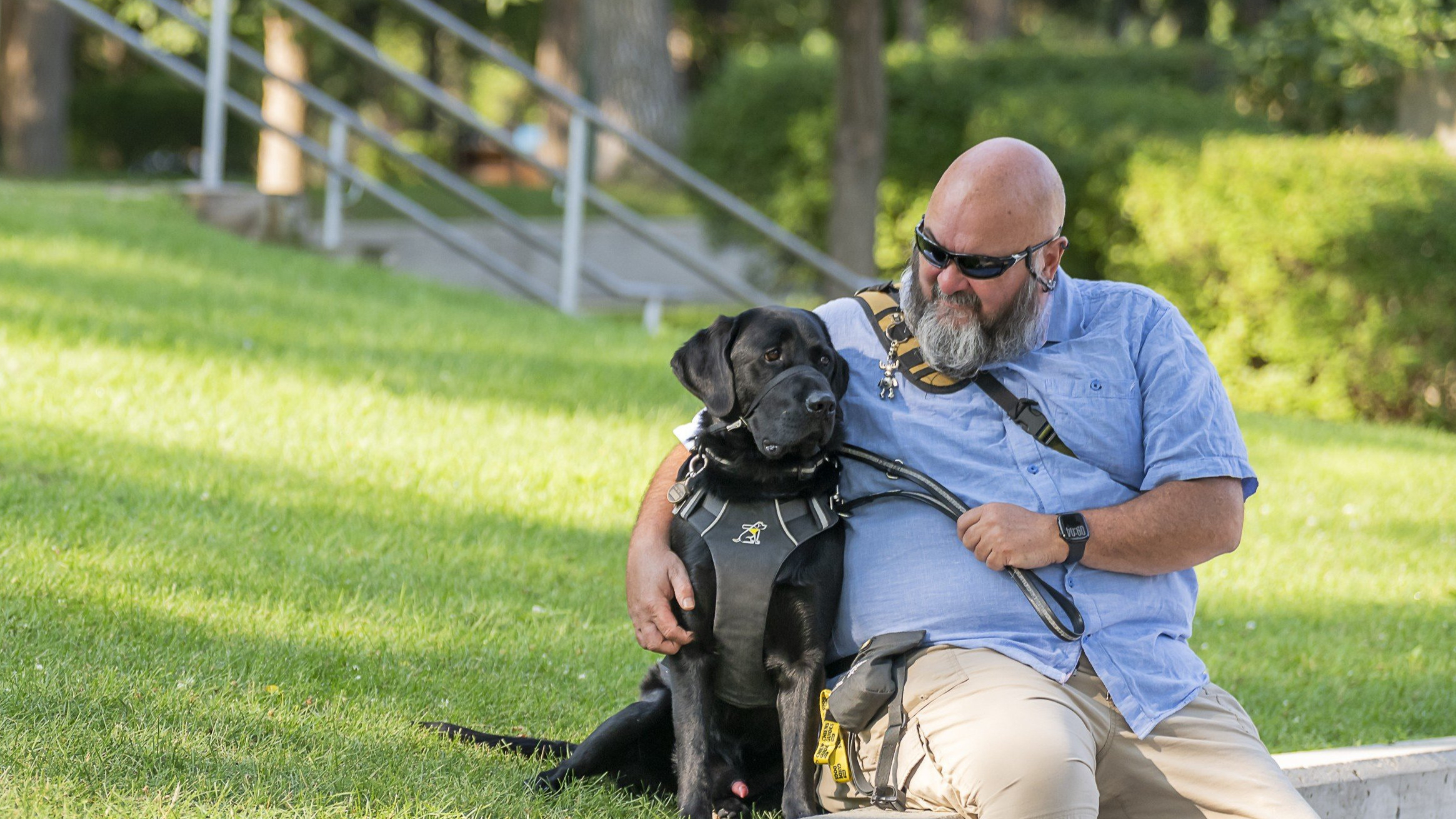 Jason sits on a park ledge beside Midas, his black guide dog. Jason is looking at Midas and smiling, and wears a light blue collared t-shirt , beige pants, and black sunglasses.