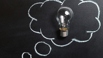 Chalkboard art. A lightbulb sits on top of a chalkboard. Surrounding the lightbulb is a chalk illustration of a thought cloud bubble. 