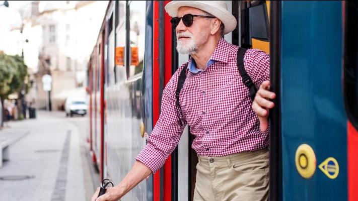 An image of an older man getting out of a streetcar and exiting onto a busy street. He uses a white cane, and is wearing a red and blue collared shirt, khakis and a fedora. He is smiling.