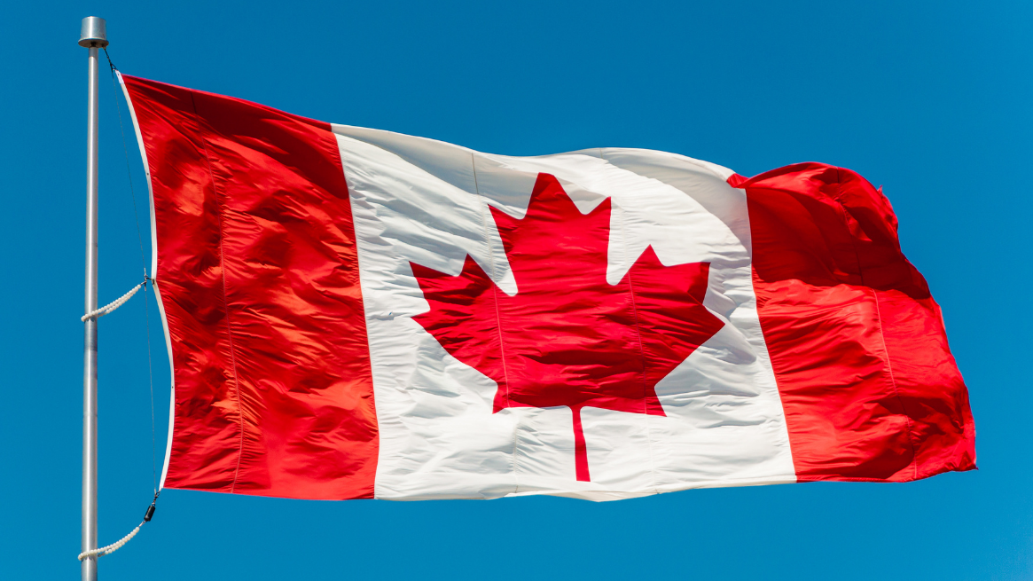 A Canadian flag waves on a flagpole against a bright blue sky.