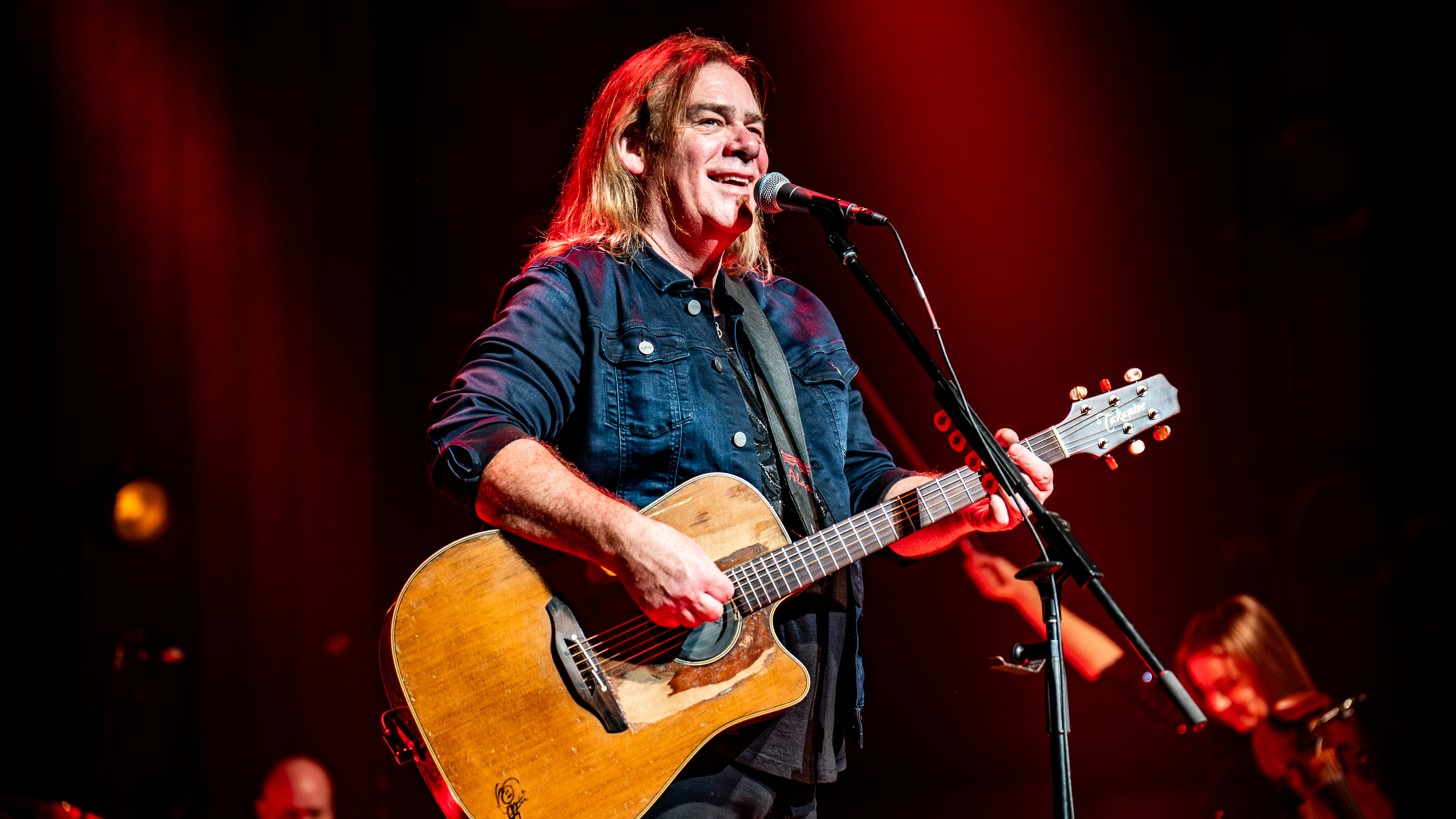 Musician Alan Doyle stands on stage in front of a microphone holding a guitar 