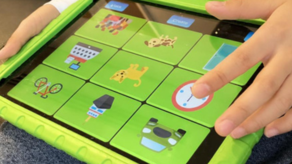 A child’s hands hold an iPad in a bright green protective case, tapping a grid of colourful icons including animals, objects, and a clock on the screen.