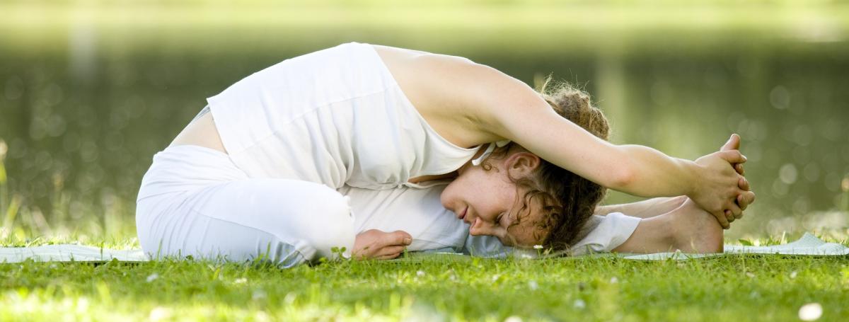 Woman in white top and pants is sitting on the grass, stretching to hold her foot with her hands.