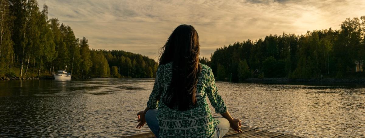Woman sitting cross-legged on a dock facing a lake and trees.