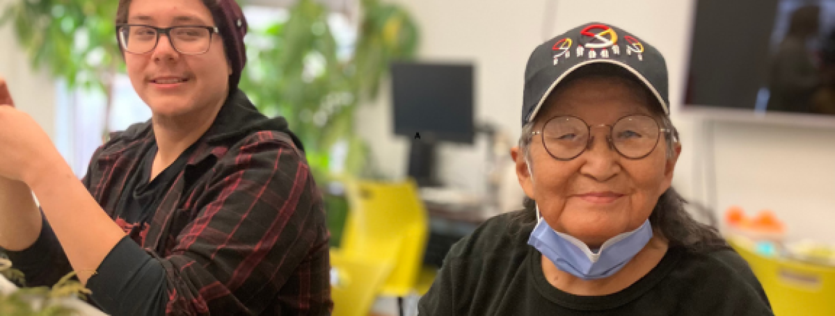 Two Indigenous participants sit at a table in CNIB Thunder Bay smiling. During a holiday program, they create decorations out of fresh cedar bows, dried orange slices, homemade gingerbread cookies, ribbons, bells, cinnamon sticks, and jute twine.