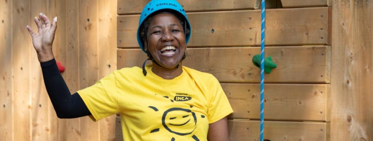 A black woman is standing in front of the climbing tower, smiling and waving with one hand. She is wearing a blue helmet and a yellow CNIB tshirt with a smiley face.