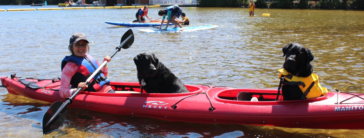 A woman is sitting in a kayak holding a paddle. There are two black guide dogs with her in the boat.
