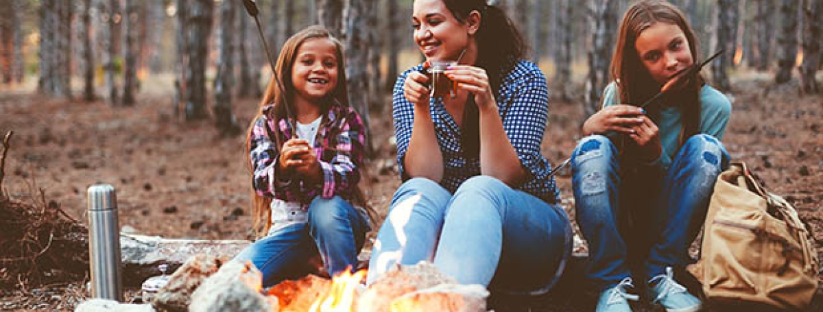 A woman and two girls sit outside in front of a fire eating treats.