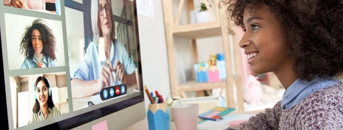 A young girl is sitting at a desk, looking at a computer screen with other people on screen.
