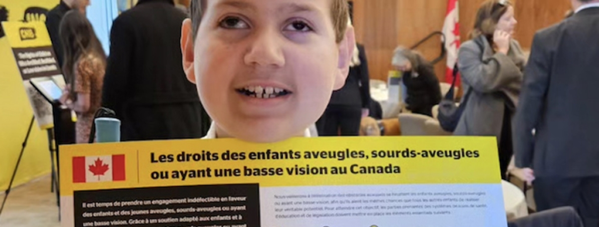 A boy is standing in a meeting room with business people behind him. He is holding up the Rights of Children Who Are blind in Canada (French version).