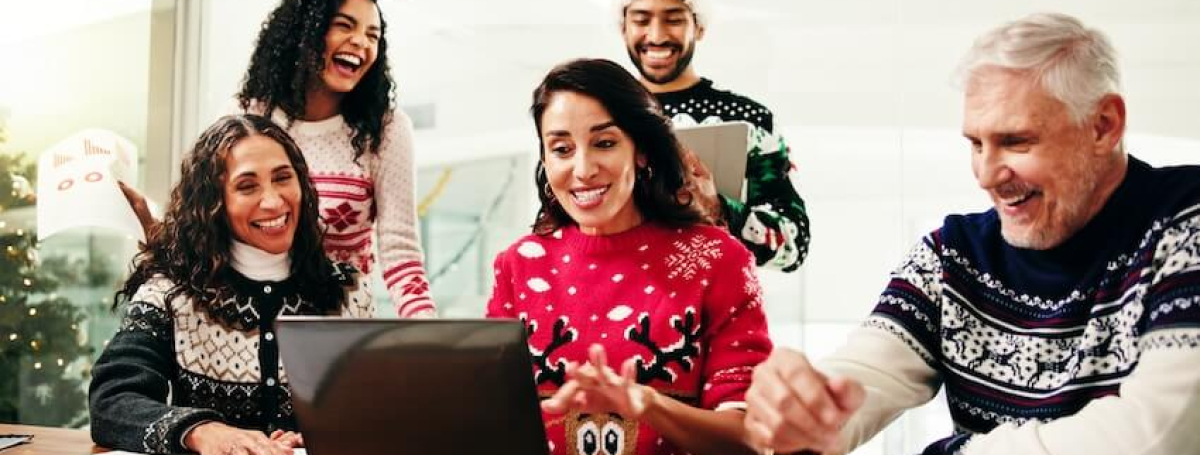 Four men and women wearing holiday sweaters and Santa hats sit around a laptop.