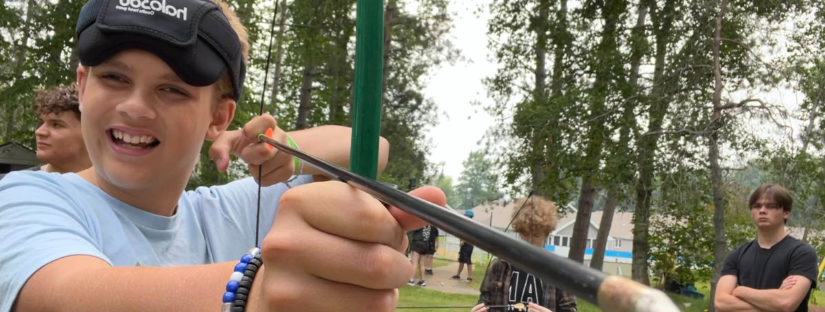 Young camper with blindfold is drawing a bow and arrow.