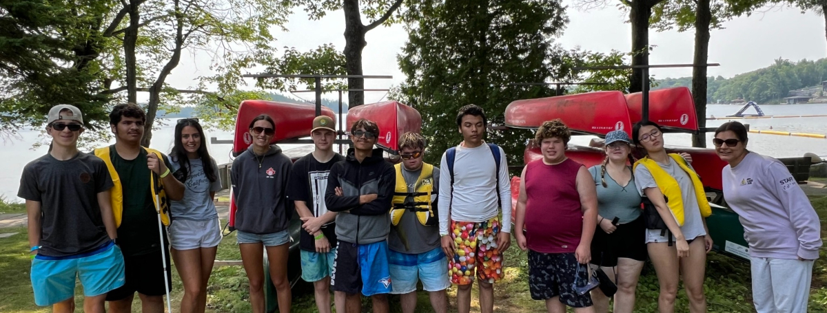 A group of youth are standing outside in front of stacked canoes. 