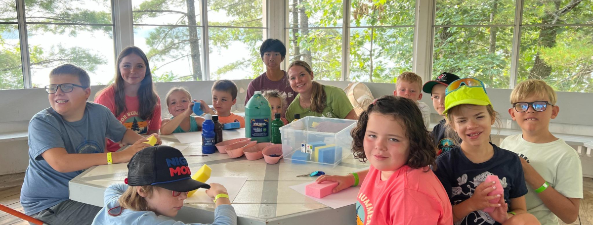 Children sitting in a gazebo at a wooden table covered with craft supplies.