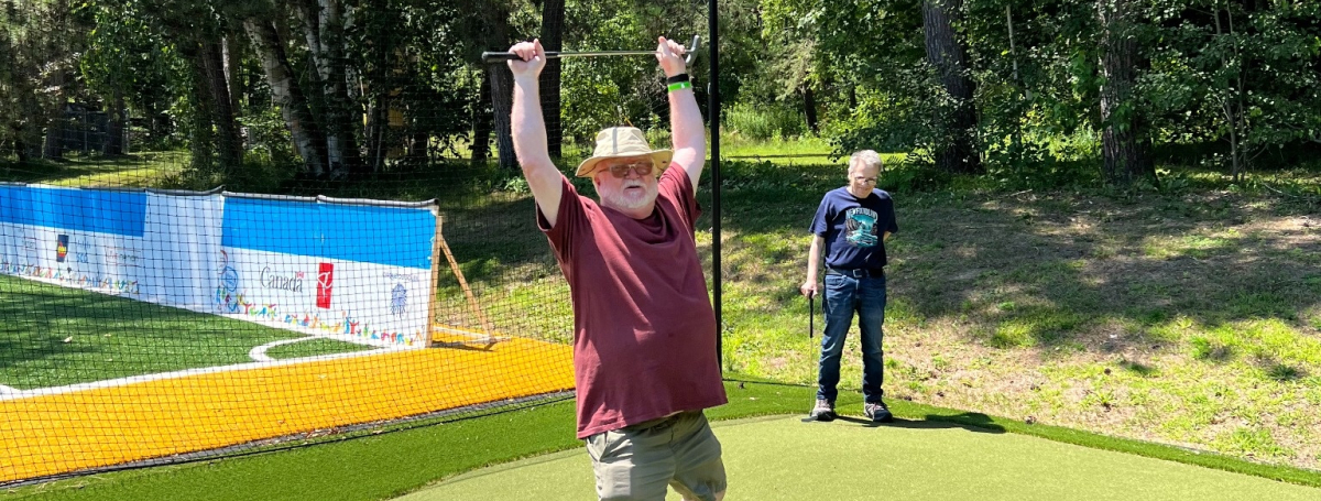 A man is standing in a mini putt course triumphantly holding a club over his head and smiling.