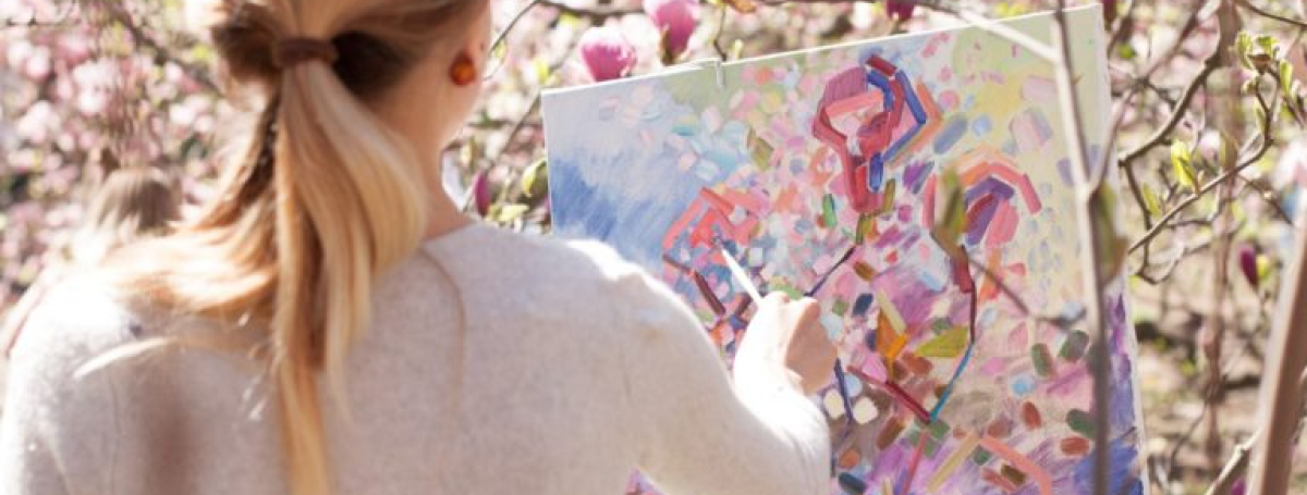 A woman is standing outside amongst bushes with blossoms. She is painting on a canvas.