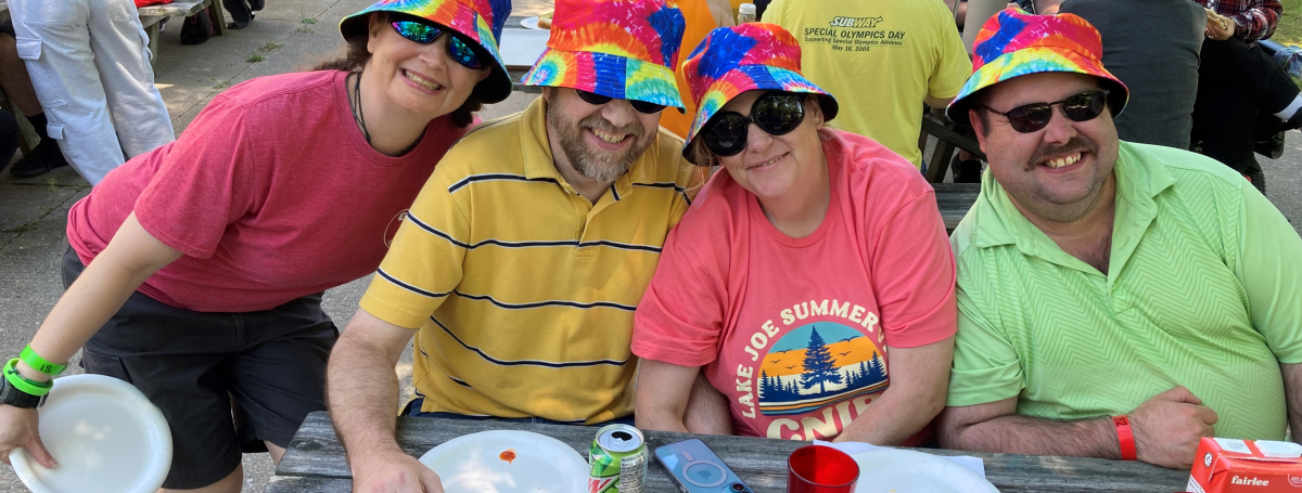 Four adults sitting outside at a picnic table wearing colourful tshirts and tye die bucket hats