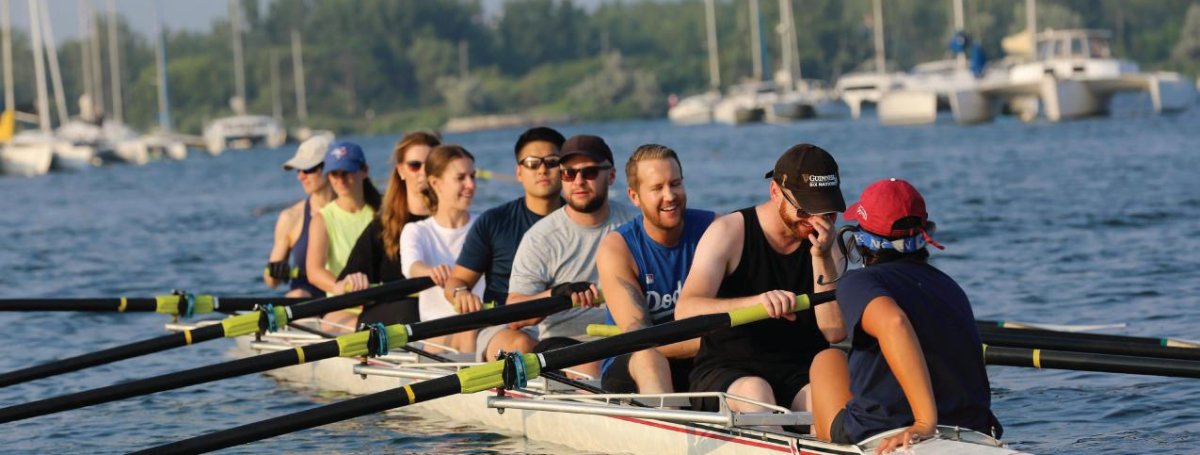A group of eight people sit in a long rowing shell on calm water, holding oars in position. They appear relaxed and smiling, as if pausing between strokes. In the background, several sailboats are anchored near a tree-lined shore.