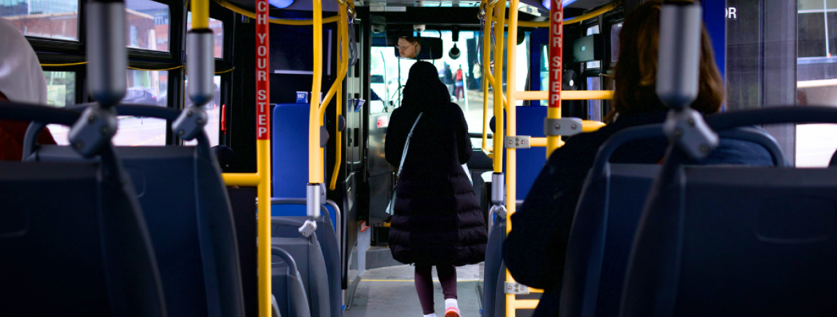 The interior of a city bus. The image is taken from the back of the bus, where a person is walking down the aisle towards the front door.
