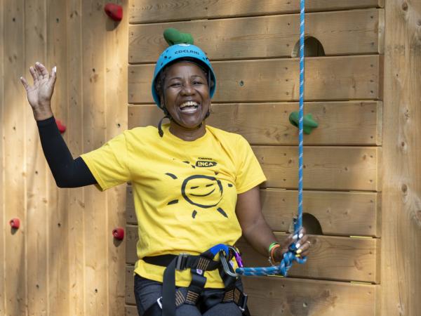 Smiling woman in a bright yellow shirt and helmet, joyfully waving, sits harnessed in front of a wooden climbing wall with colorful handholds.