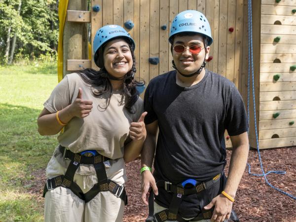 Two people wearing helmets and harnesses stand in front of an outdoor climbing wall, smiling at the camera; one gives a thumbs up.