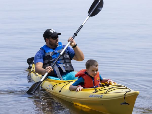 An adult and a child wearing life jackets paddle a yellow kayak together on calm water.