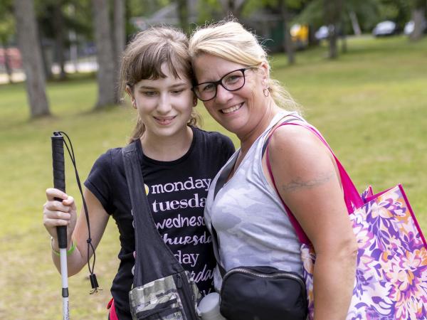 A young girl with a white cane and a woman smiling together in a park, surrounded by trees and grass.