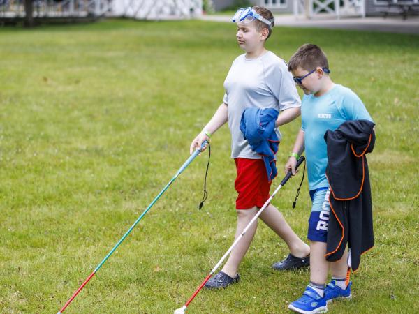 Two boys walking on grass, each holding a white cane and wearing swim gear, with one boy in red shorts and the other in blue.