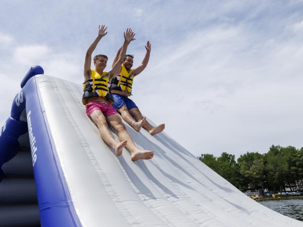 Two young men in life jackets sliding down an inflatable water slide into a lake, with trees and a cloudy sky in the background.