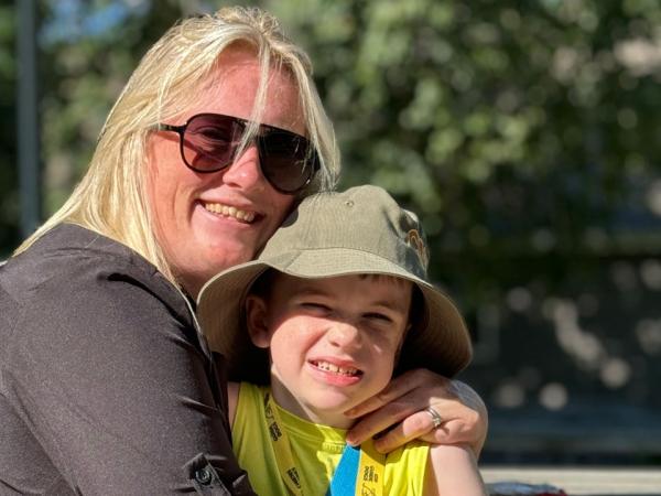 A woman with blonde hair wearing sunglasses smiles while holding a young boy in a yellow shirt and a hat.