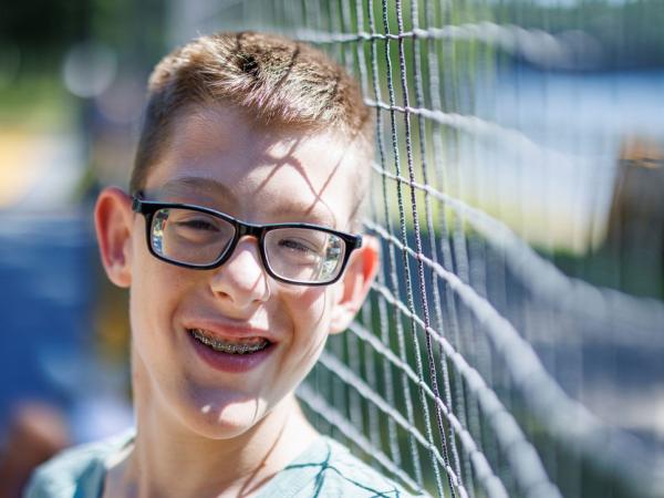 Smiling boy with glasses and braces standing near a fence, wearing a light green shirt, with a blurred background of trees and water.