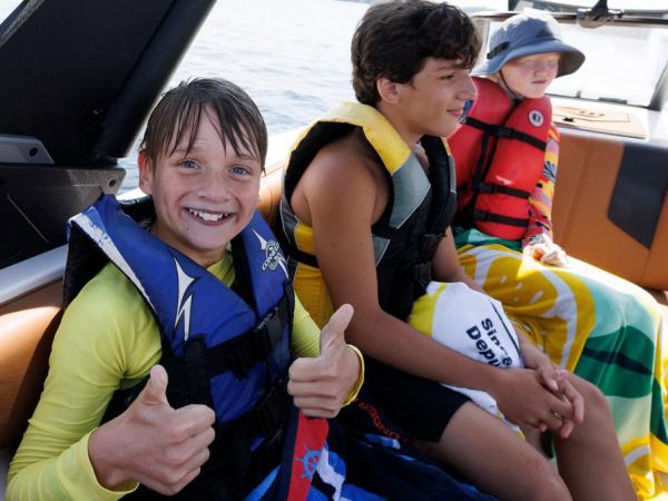 Three children sitting on a boat, wearing life jackets and smiling. One child is giving a thumbs-up gesture.
