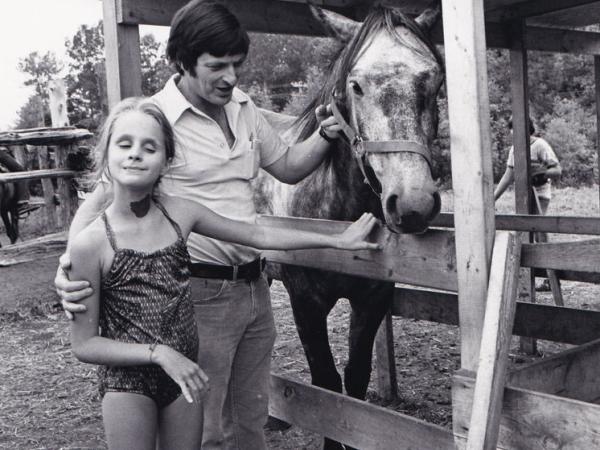 Man and young girl pet a horse in a fenced area. The girl smiles contentedly with closed eyes, while the man gently holds the horse’s bridle. Rustic setting.