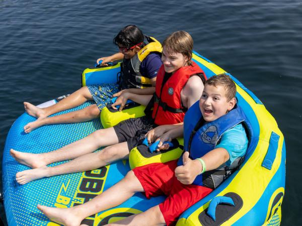 Three boys wearing life vests sit on a colorful inflatable tube on water. They are smiling and appear excited, capturing a playful, adventurous mood.