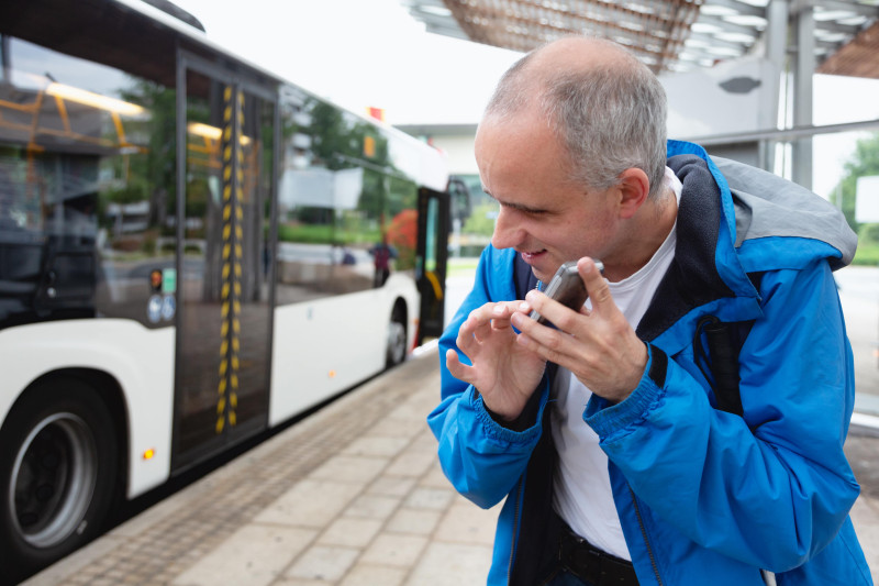 A man stands by a bus touching his mobile phone and listening to it.