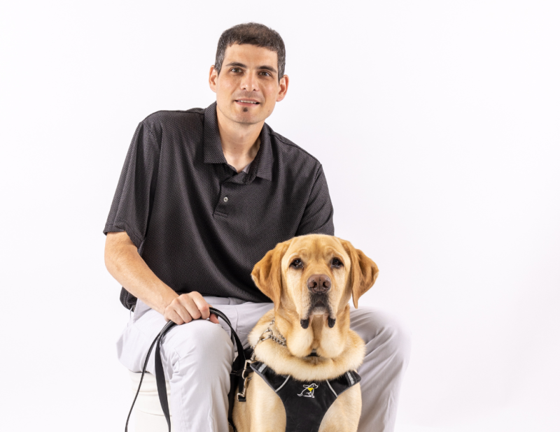 Ryan, a guide dog handler, is seated. Ryan's guide dog, a yellow-lab named Joe, is in harness/on leash and sits in-between Ryan's legs 