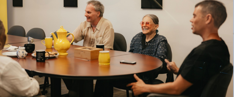 Three people having a meeting at a table, there are mobile phones and a yellow tea pot on the desk