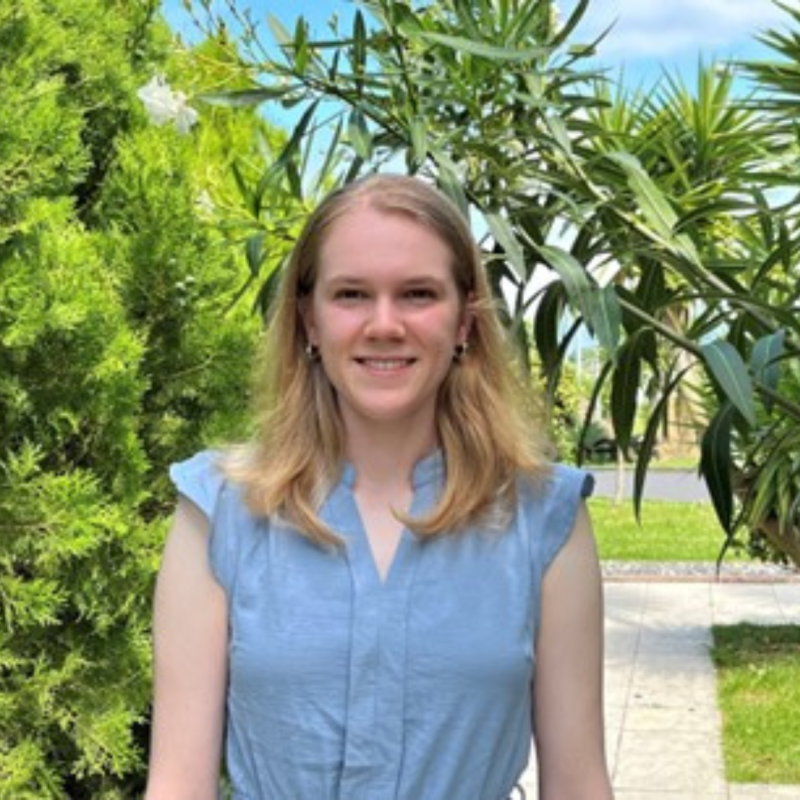 Laura, a young woman with blonde hair, smiles while standing in front of a lush green garden with palm trees and shrubs. She is wearing a light blue sundress. 