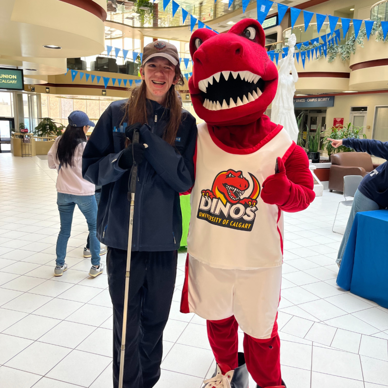 Zachary, a young man with long brown hair, smiles while standing next to the university of Calgary's fluffy red dinosaur mascot, Rex. Zach is wearing a blue sweatshirt, sweatpants, a hat, and holds his white cane. 