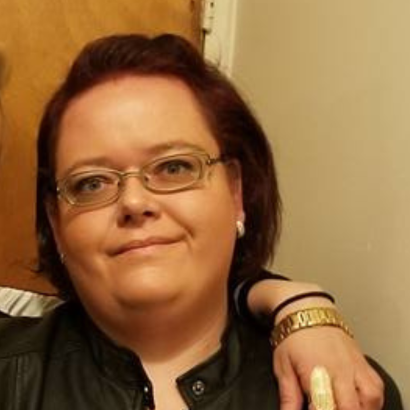 Jaclyn, a young woman with red hair and glasses, smiles and poses for a selfie-style photograph. 