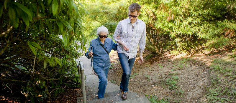 An older woman who is holding a white cane and wearing sunglasses walks with a younger man down a forest path
